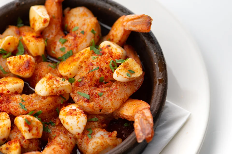 Photo d’illustration : Crevettes à l'ail garnies d'herbes hachées, servies dans un bol noir posé sur une assiette blanche